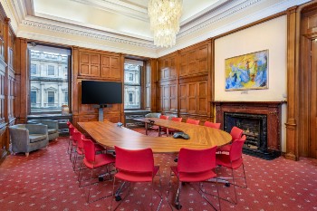 gertrude elles meeting room with wood panelled walls, a fireplace, two sash windows and an oval table with red chairs around it